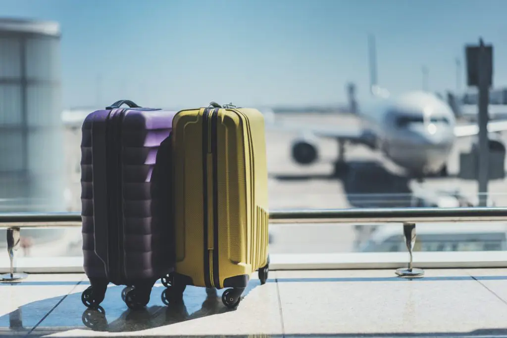 A purple suitcase and a yellow suitcase by the window overlooking airside with a plane parked in front