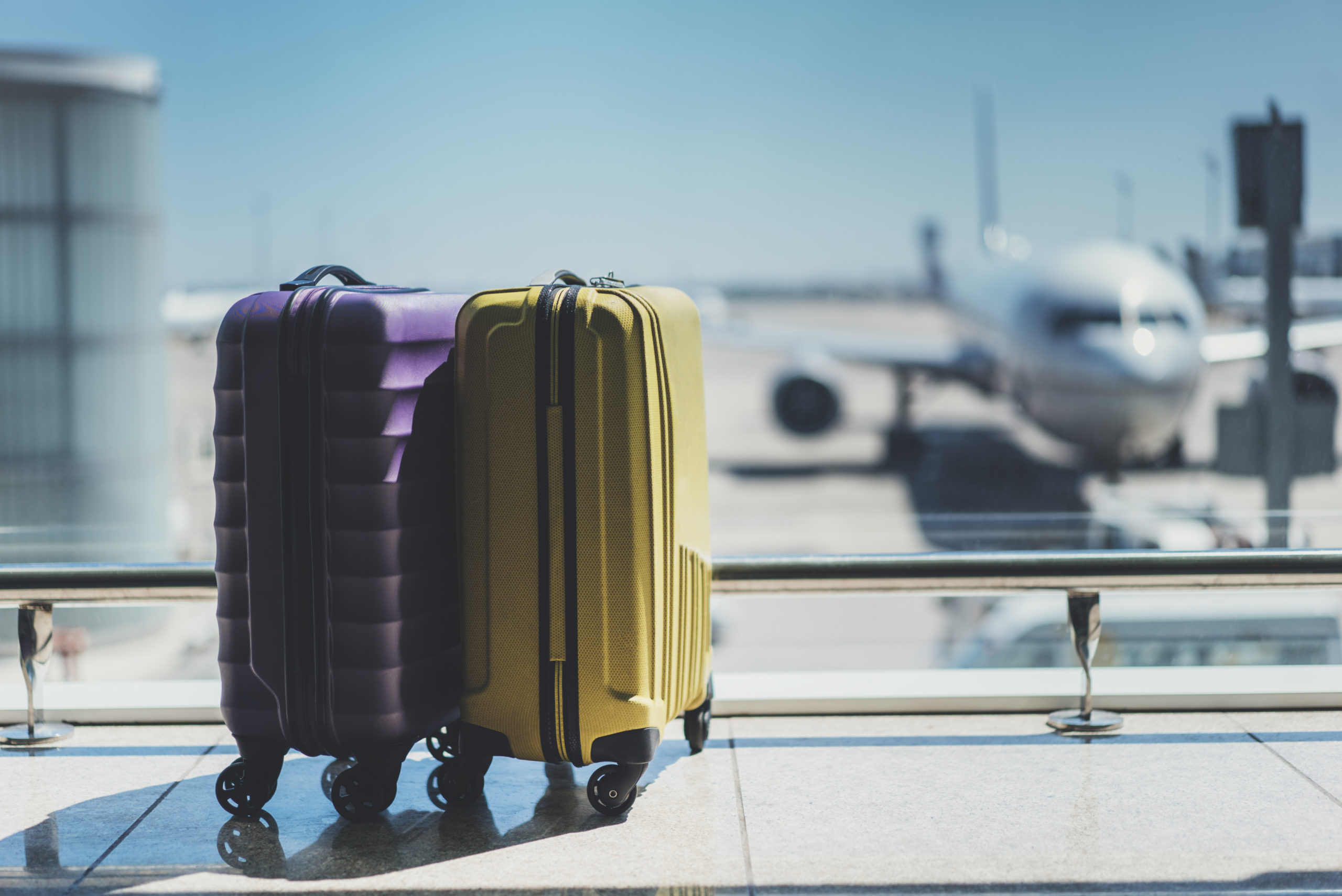 Une valise violette et une autre jaune devant la fenêtre donnant sur la piste avec un avion devant.