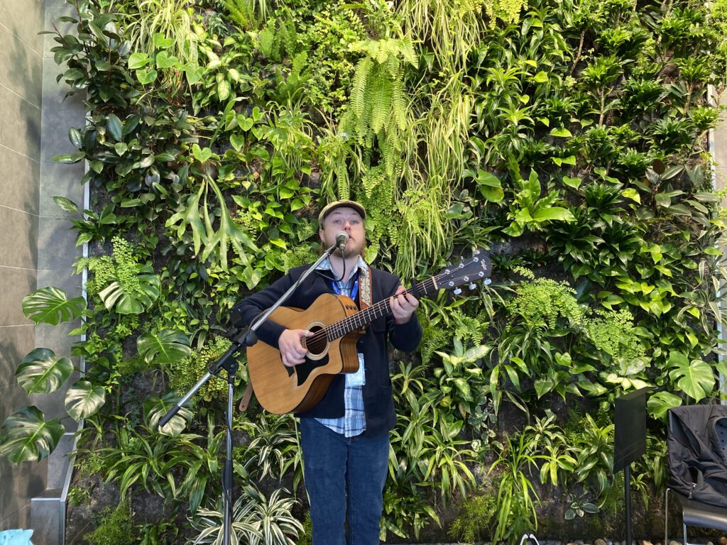 Musician with guitar singing into the microphone with greenery in the background