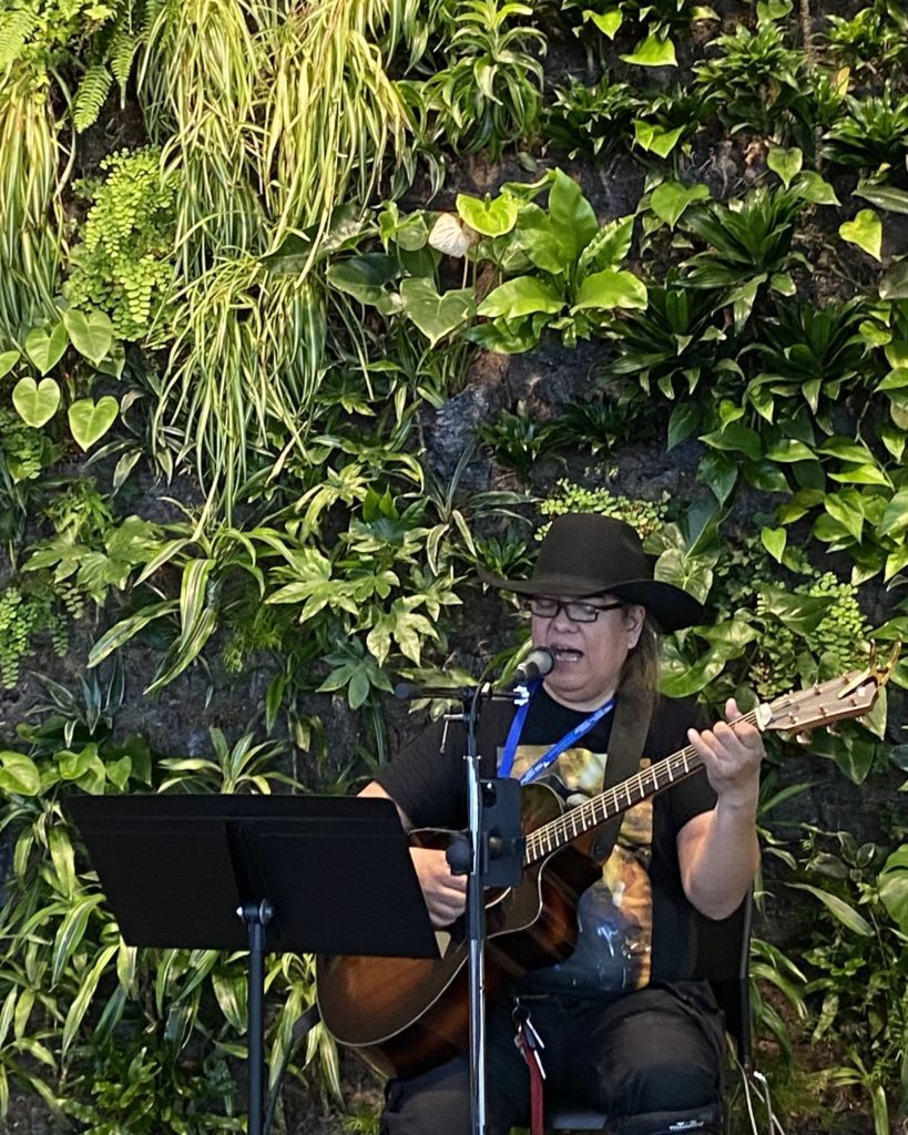 Musician in cowboy hat playing guitar and singing into microphone in front of a wall filled with green plants